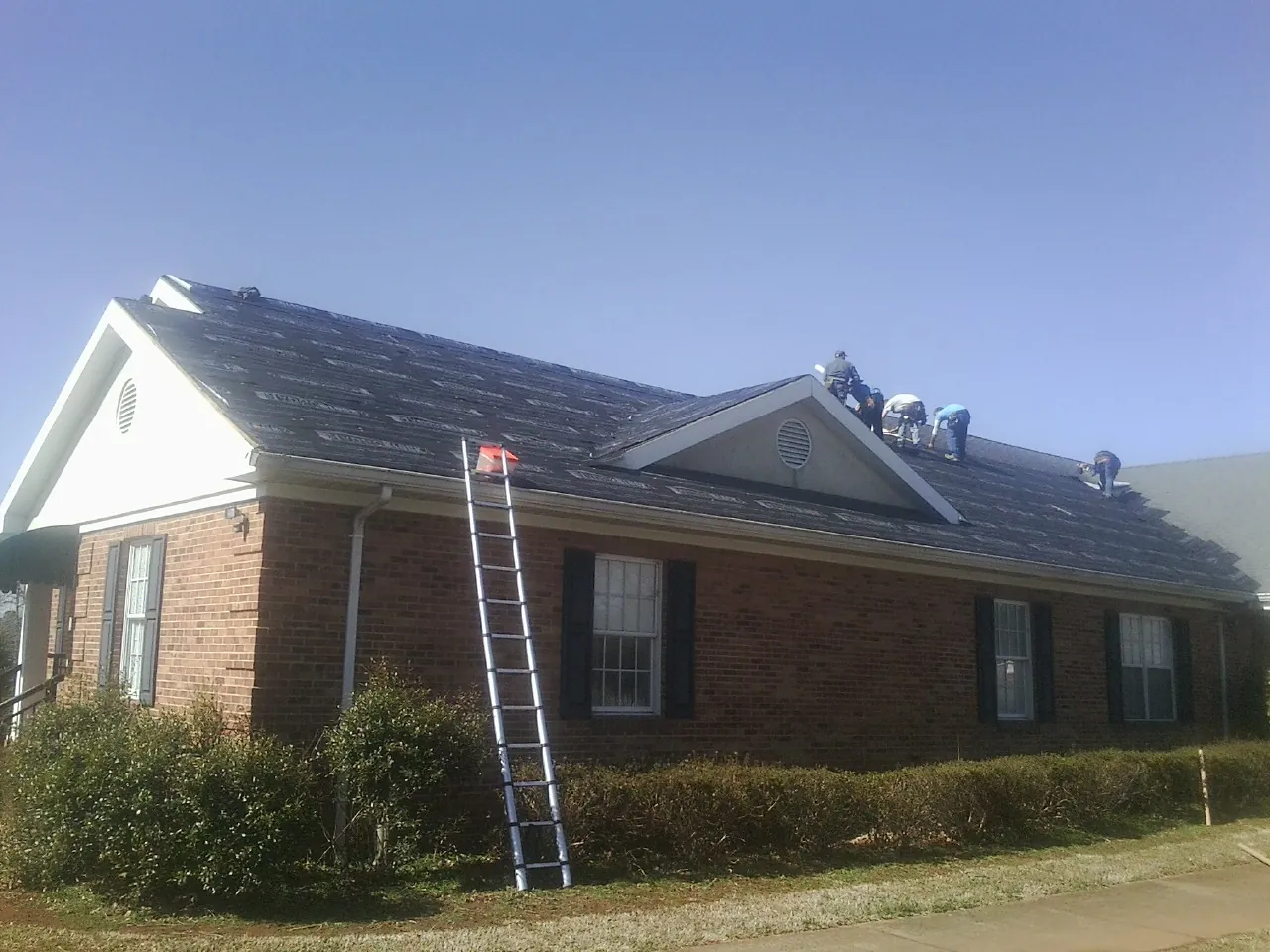 Crew tearing shingles off a brick ranch roof with a ladder set — Metal Roof Repair work by our crew in Elderton