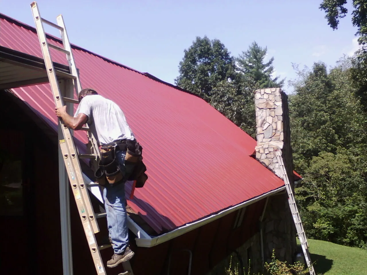 Roofer on a ladder beside a red metal roof and stone chimney — Soffit & Fascia Repair by Roofing Brothers in Elderton